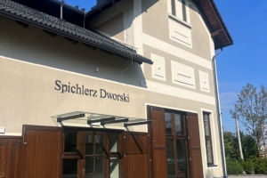 Budynek o jasnej elewacji z ciemnym dachem, dużymi drewnianymi drzwiami i przeszklonymi oknami na parterze. Przed budynkiem znajduje się kamienny podjazd, a w tle widać drzewa i niebo.